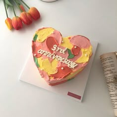 a heart shaped cake sitting on top of a table next to an open book and tulips