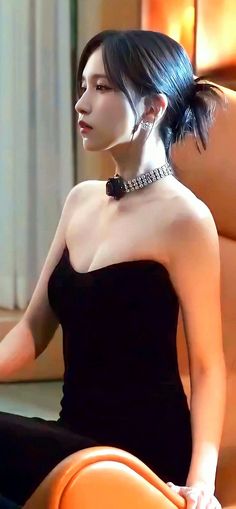 a woman sitting on an orange chair wearing a black dress and necklace with her hair in a pony tail