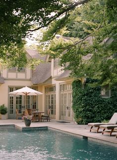 an outdoor swimming pool with lounge chairs and umbrellas in front of a house that is surrounded by trees