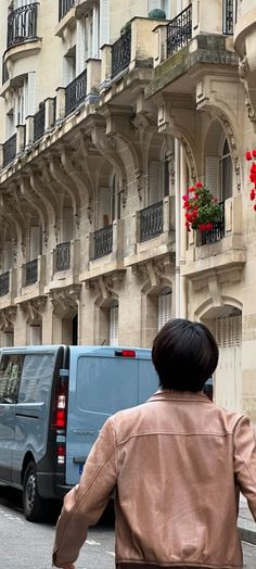 a woman walking down the street in front of a building with red flowers hanging from it's balconies