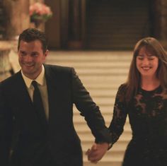 a man and woman holding hands walking down a hall way in formal wear, with flowers on the table behind them