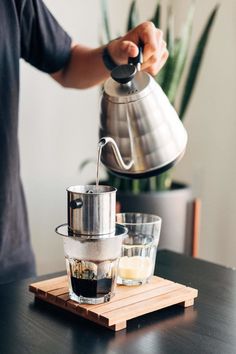 a person pours coffee into a glass cup on a wooden tray next to a potted plant