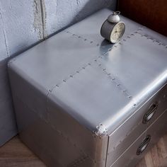 a metal box with a clock on it sitting next to a brick wall and wooden floor