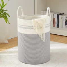 a white and grey basket sitting on top of a rug next to a potted plant