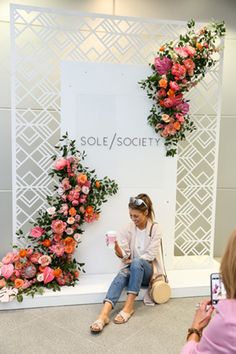 a woman sitting on the ground in front of a wall with pink flowers and greenery