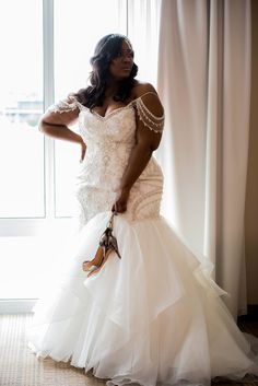 a woman in a wedding dress standing by a window with her hand on her hip