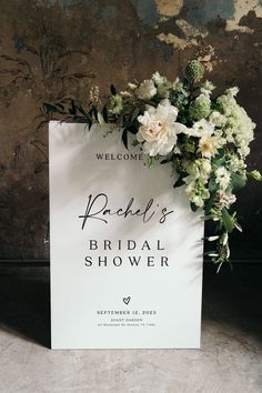 a bridal shower sign with flowers and greenery on the front, against a rusted wall