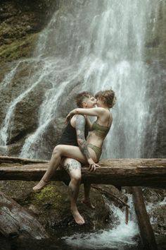 two people are sitting on a log in front of a waterfall and one is kissing the other