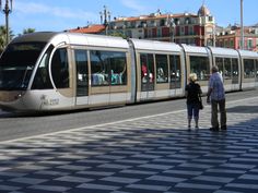 two people are standing on the sidewalk next to a train that is pulling into town