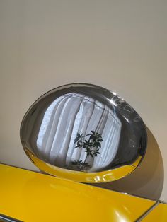 a mirror with a plant in it on top of a yellow table next to a white wall