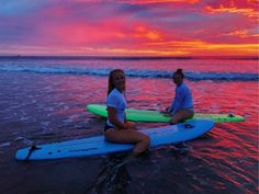 two women sitting on surfboards in the water at sunset, with one holding her leg up