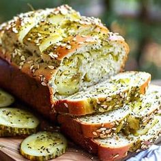 sliced loaf of bread with pesto and sesame seeds on wooden cutting board next to sliced cucumbers