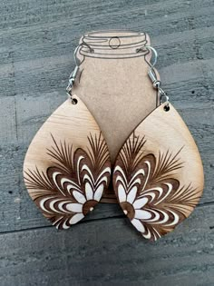 two wooden earrings with brown and white designs hanging from hooks on a wood floor next to a mason jar