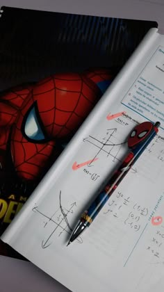 an open book with a pen on top of it next to a spiderman drawing