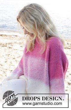 a woman sitting on the beach wearing a pink and white sweater