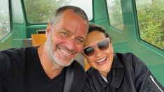 a man and woman taking a selfie in front of a green vehicle with trees behind them