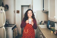 a woman standing in a kitchen holding a cup and smiling at the camera while wearing an apron