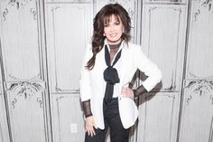 a woman standing in front of a wall wearing a white jacket and black tie with her hands on her hips