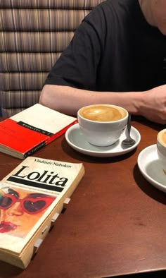 a man sitting at a table with two cups of coffee in front of him and a book