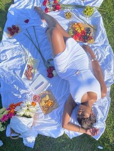 a woman laying on top of a white blanket next to fruit and flowers in the grass