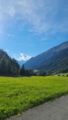 an empty road in the middle of a grassy field with mountains in the background on a sunny day