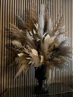 a vase filled with lots of dry grass on top of a glass table next to a striped wall