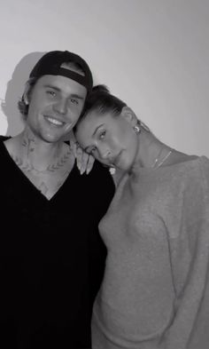 a man standing next to a woman in front of a white wall wearing a baseball cap