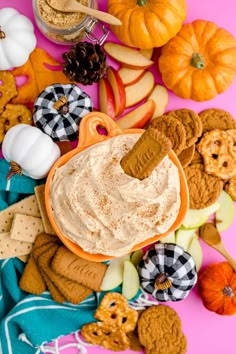 a bowl of peanut butter surrounded by cookies, crackers and pumpkins on a pink background