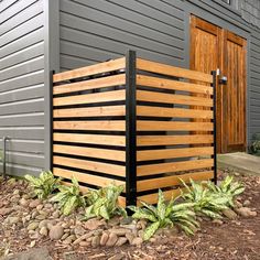 a wooden fence in front of a gray building with green plants and rocks on the ground