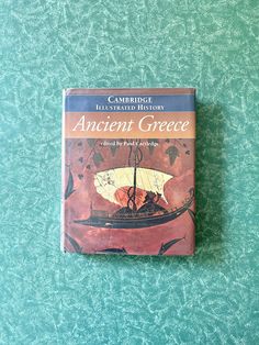 the book ancient greece is sitting on top of a blue tablecloth covered surface with an image of a boat in the water