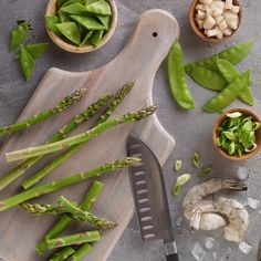 asparagus and other vegetables on a cutting board next to a knife, bowl with shrimp