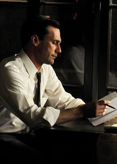 a man sitting at a table with a pen and paper in his hand while looking out the window