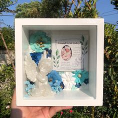 a hand holding up a white box with flowers and butterflies in the center, on top of a garden