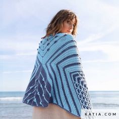 a woman standing on the beach wearing a blue knitted shawl