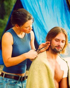 a man getting his hair cut by a woman in front of a blue tarp