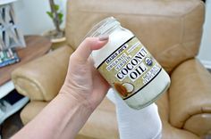 a hand holding a bottle of coconut oil in front of a brown leather chair and coffee table