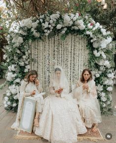 two brides sitting in front of a floral arch with flowers on the wall and white curtains behind them