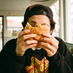 a young man eating a large sandwich in front of his face and wearing a black hoodie