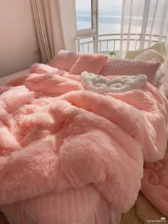 a bed covered in pink fluffy blankets next to a window with a view of the ocean