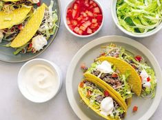 three tacos and two bowls of salad on a white tablecloth with condiments