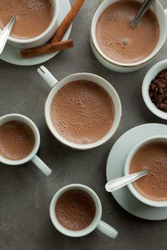 four cups of hot chocolate with spoons and cinnamon on the side, surrounded by other mugs