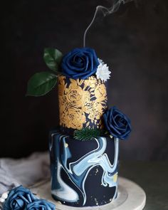 a three tiered cake with blue roses on top and gold leaf decoration, sitting on a white plate