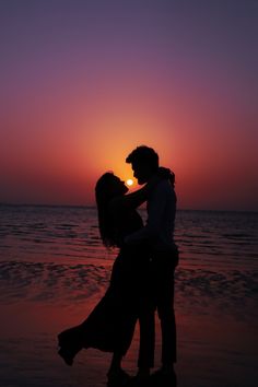 a man and woman kissing on the beach at sunset with the sun setting in the background