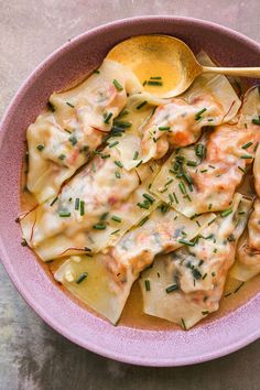 a pink bowl filled with pasta covered in sauce and herbs next to a wooden spoon