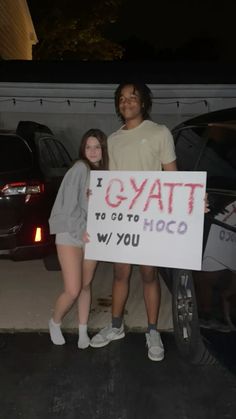 two people standing next to each other in front of a car holding a sign that says gayt to do hoo w / you