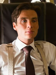 a man wearing suspenders and a white shirt is sitting in an airplane looking at the camera