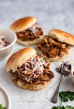 three pulled pork sandwiches with cole slaw on the side and two small white bowls