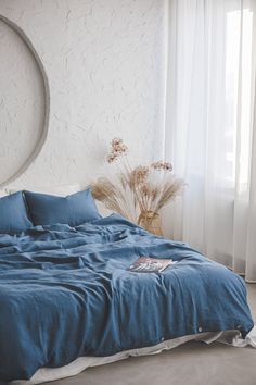 a bed with blue sheets and pillows next to a round mirror in a white room