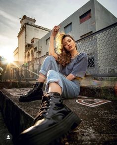 a woman sitting on the ground with her legs crossed and wearing black boots, in front of an industrial building