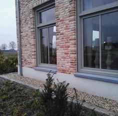 two windows on the side of a brick building with gravel and bushes in front of them